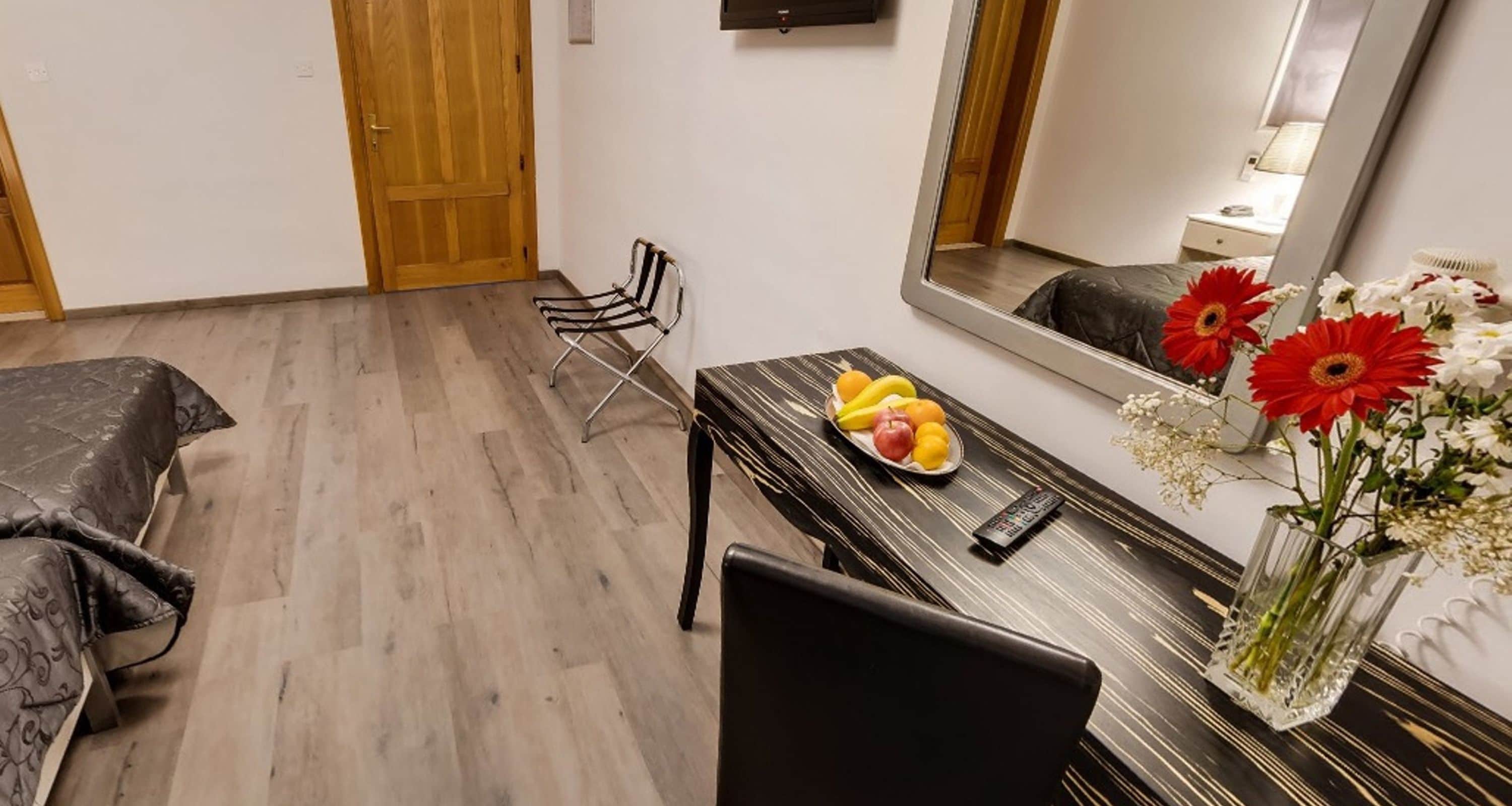 A hotel room with a bed, a dining table with fruit and flowers, a chair, and a large mirror.