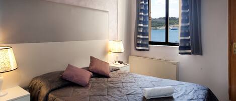 A neatly made bed with a patterned bedspread, two pillows, and a rolled towel. There are two bedside lamps, a nightstand, and a window with curtains.