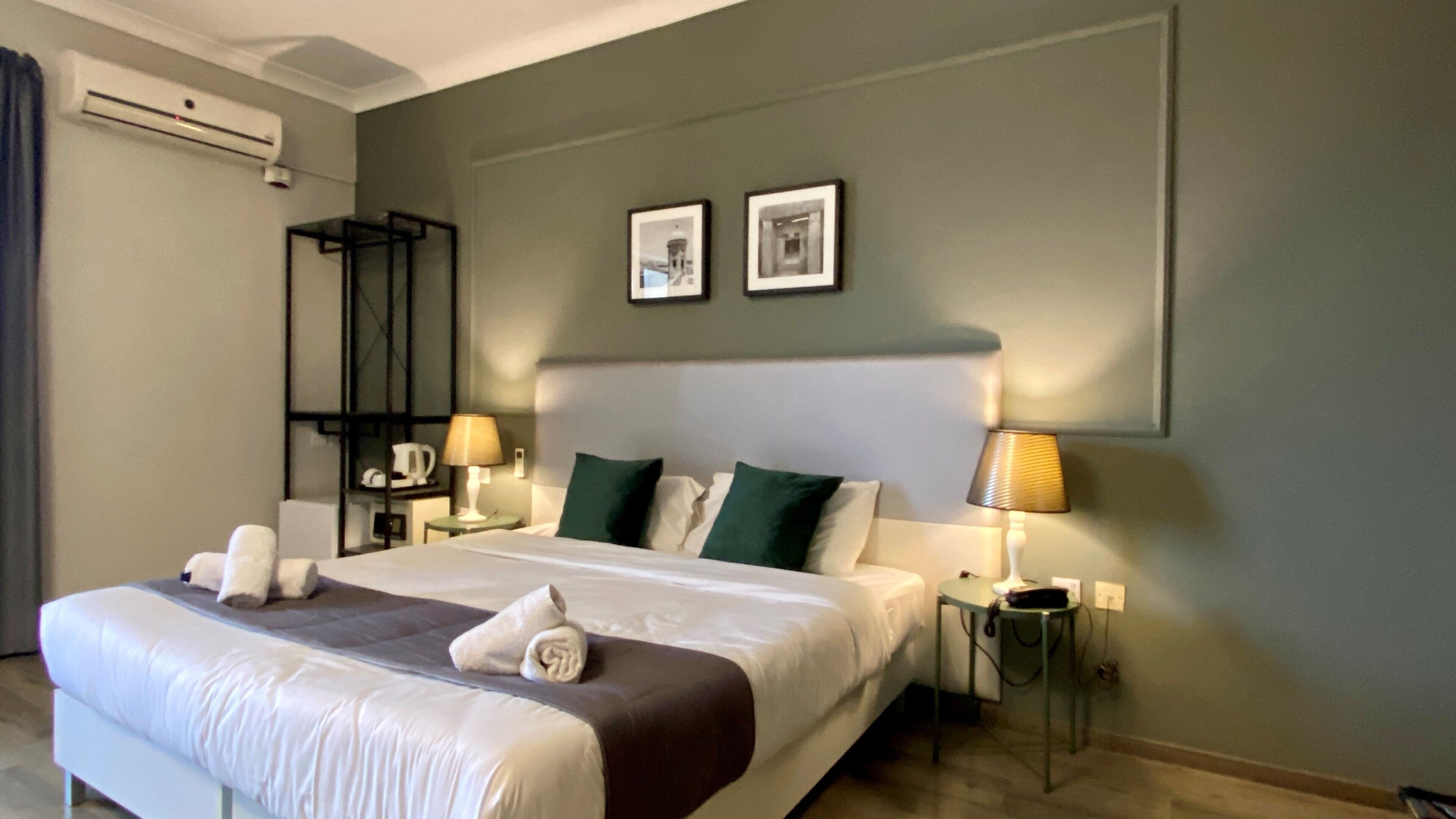 Una camera d'albergo con un letto grande, due comodini con lampade, un condizionatore d'aria a muro e due quadri incorniciati sopra il letto.