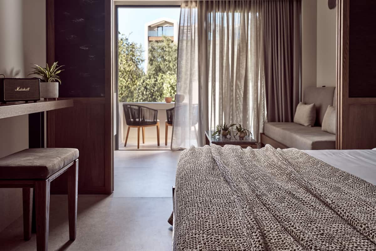 Et hotelværelse med seng, sofa, skrivebord med radio, stol, bord med planter og udsigt til det fri gennem et vindue med gardiner.