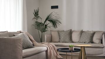 Una sala moderna con un sofá color beige, cojines estampados, una mesita redonda con una planta y una planta verde alta al lado del sofá.