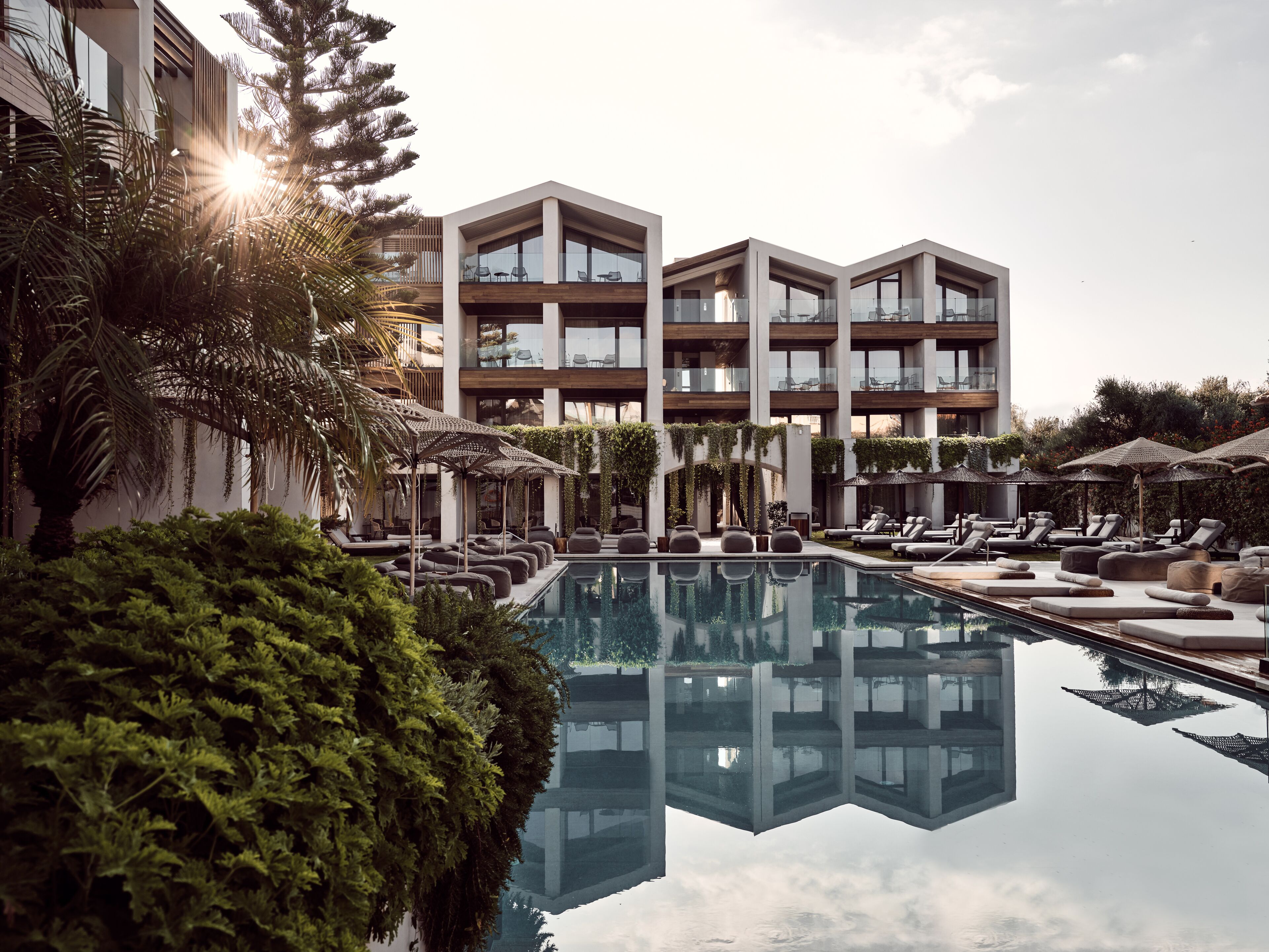 Un hôtel moderne doté d’une piscine, de chaises longues et dont l’image se reflète dans l’eau.