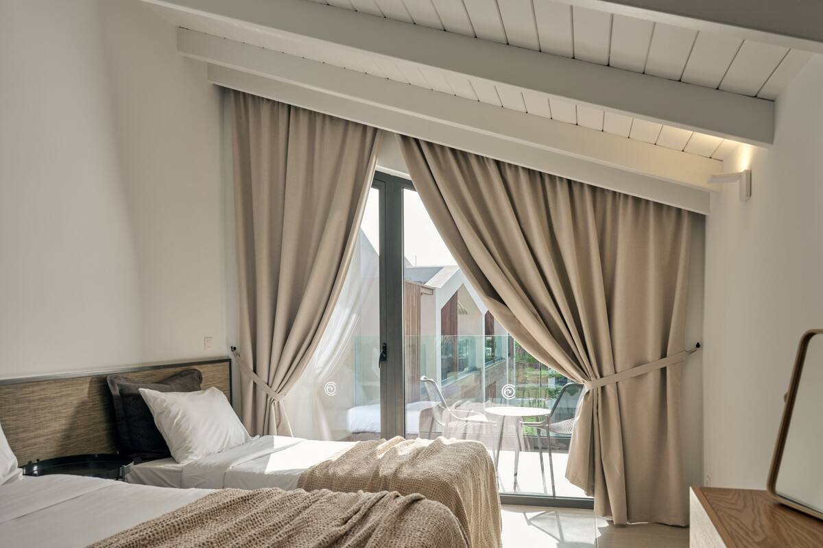 Et hotelværelse med to senge, en balkon med bord og stole, samt store vinduer med gardiner.