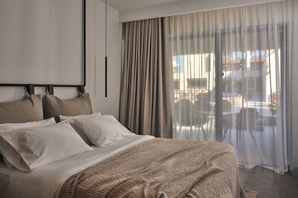 Et moderne hotelværelse med en stor seng, en sengegavl med mange puder, en hængende pendel og udsigt gennem lette gardiner til et udendørs område med møbler.
