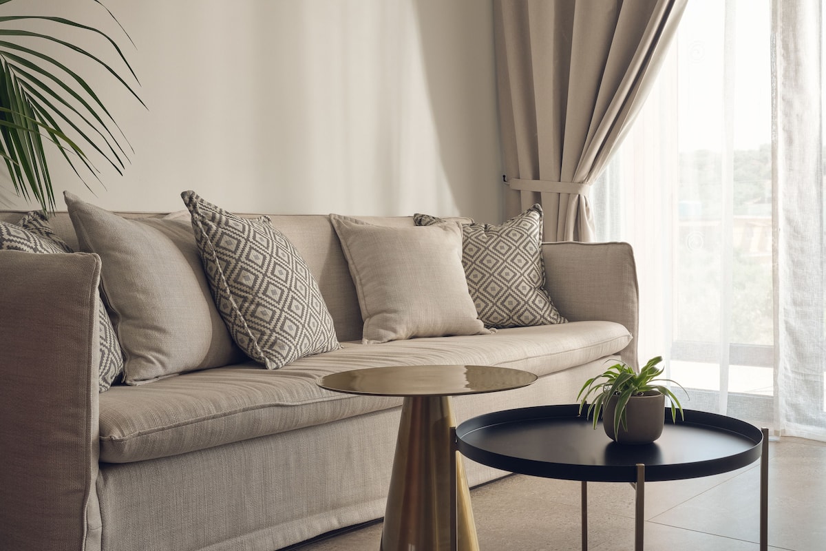 En stue med en beige sofa, mønstrede puder, et lille rundt bord med en plante og lette gardiner.