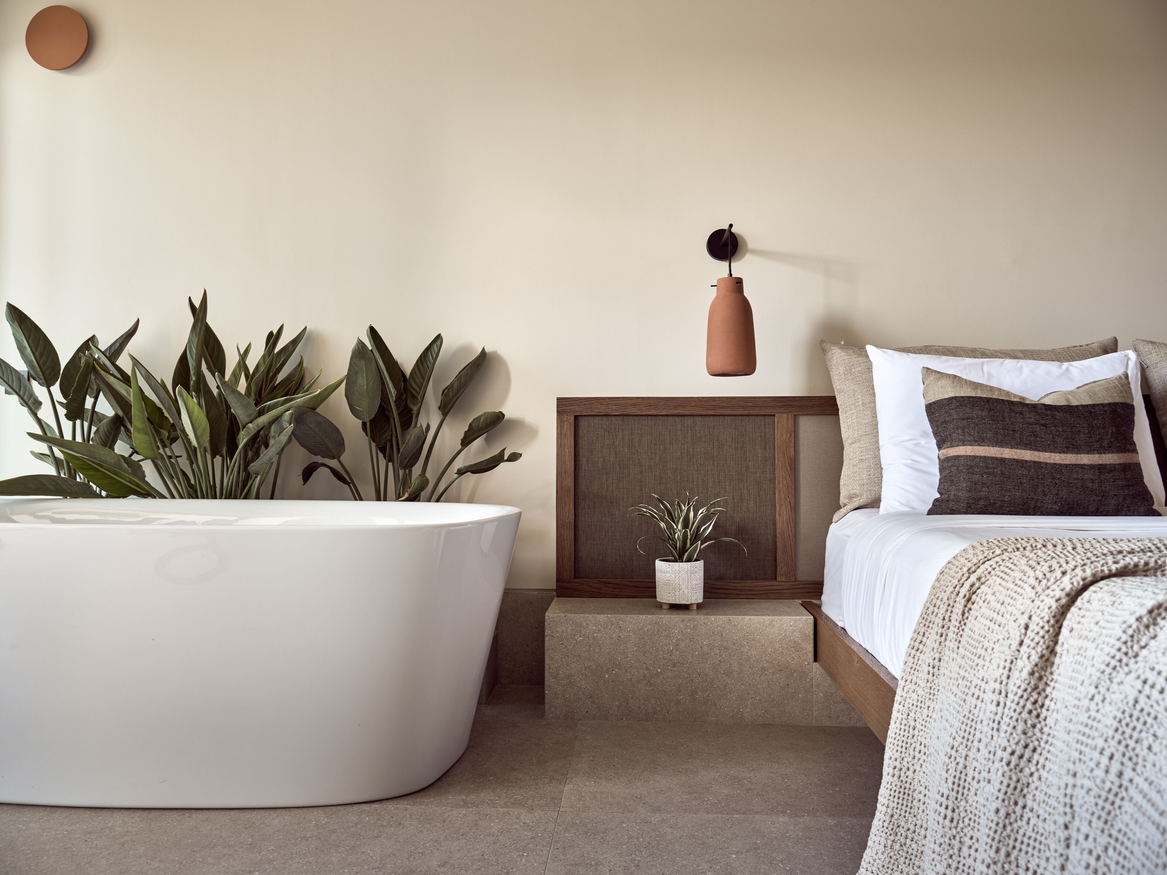 Un dormitorio moderno con una bañera blanca grande, una mesita de noche de madera con una planta pequeña y una cama con almohadas a rayas.
