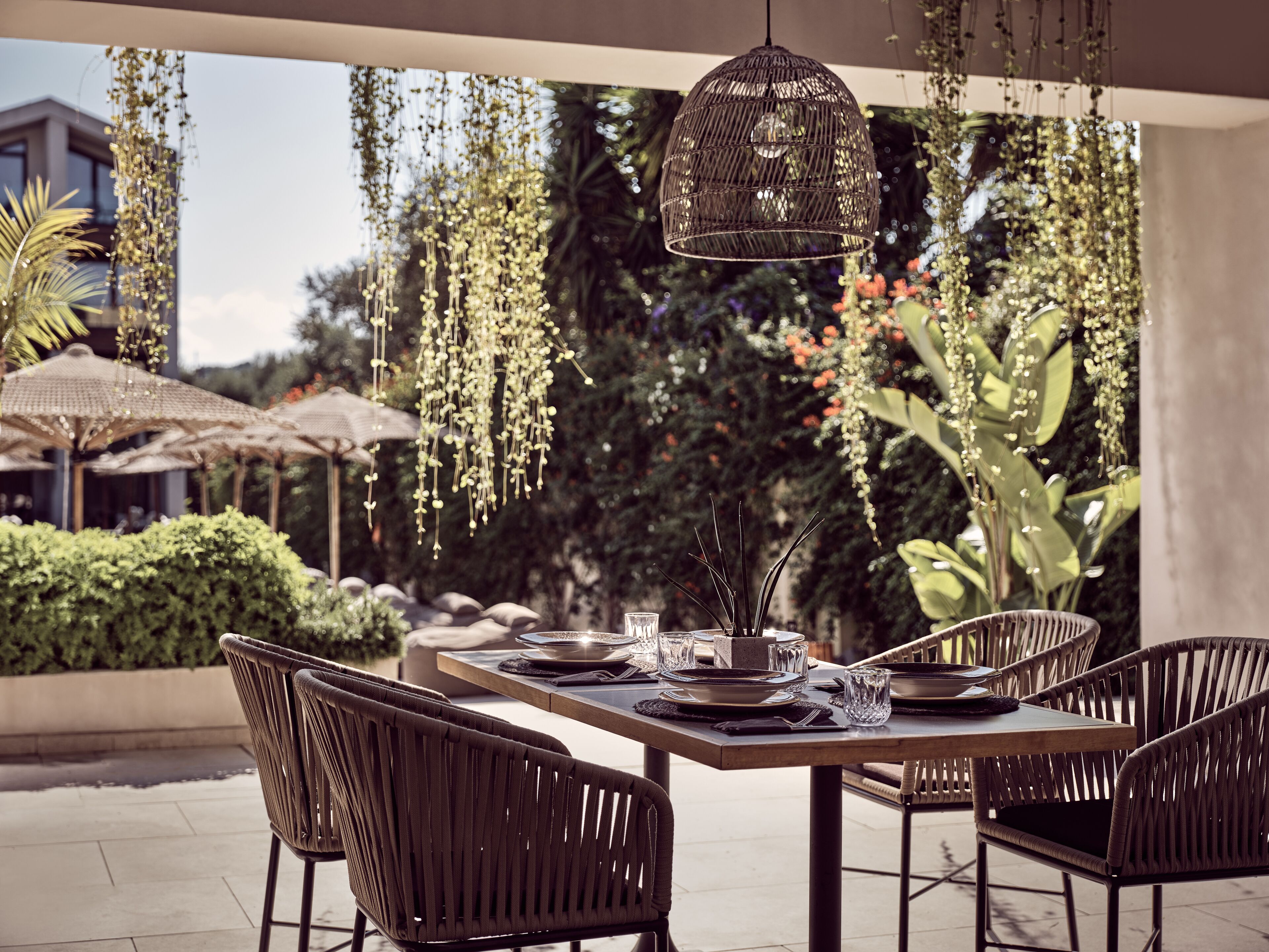 An outdoor dining area with wicker chairs and a wooden table set for two, surrounded by hanging plants and greenery.