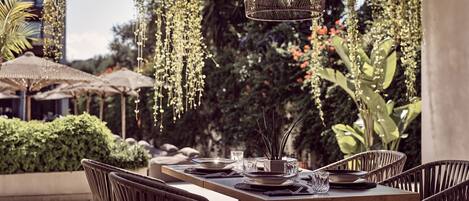An outdoor dining area with wicker chairs and a wooden table set for two, surrounded by hanging plants and greenery.
