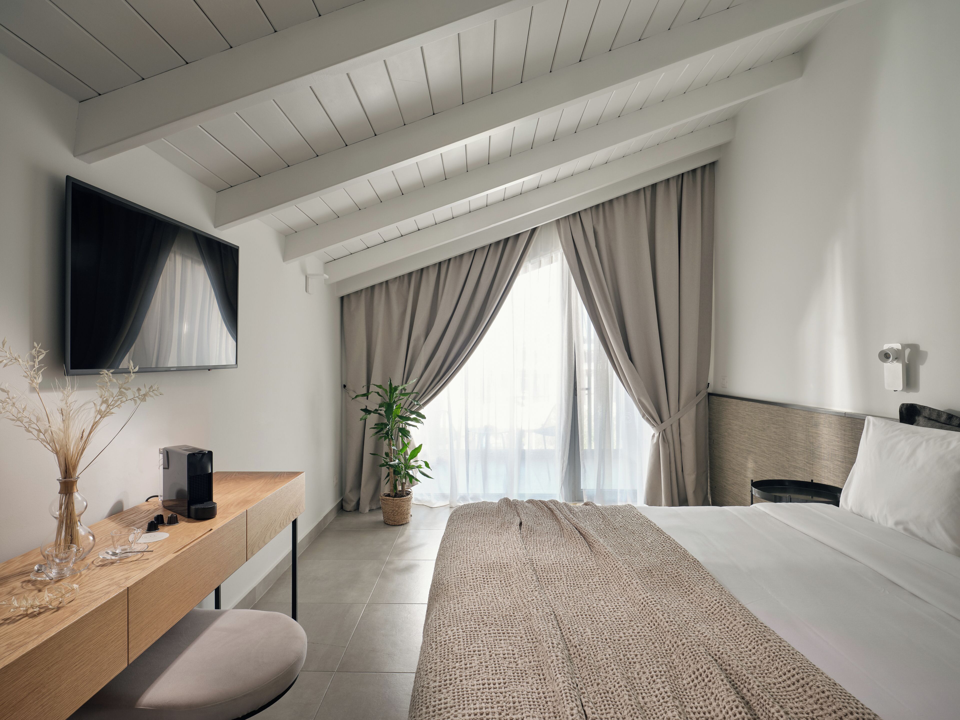 Una habitación de hotel moderna con un escritorio de madera, un televisor de pantalla plana, una cama con una manta texturizada, una planta en maceta y un amplio ventanal con cortinas.