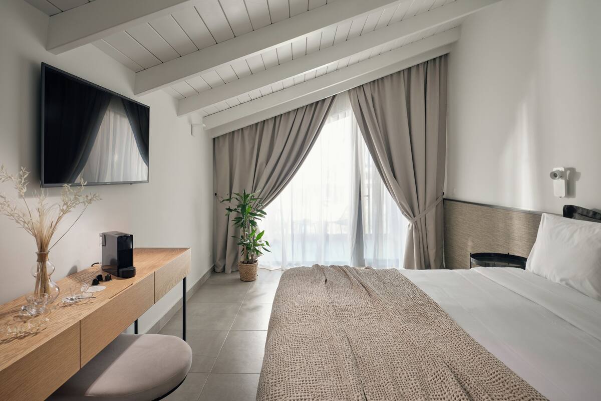 Et moderne hotelværelse med et træskrivebord, et fladskærms-tv, en seng med et vamset tæppe, en potteplante og et stort vindue med gardiner.