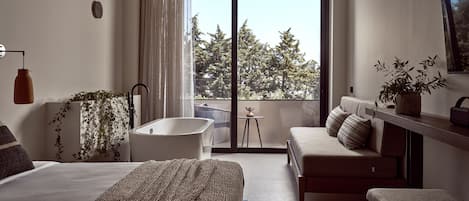 A modern bedroom with a large bed, a sofa, a small table, and a view of trees through a sliding glass door.