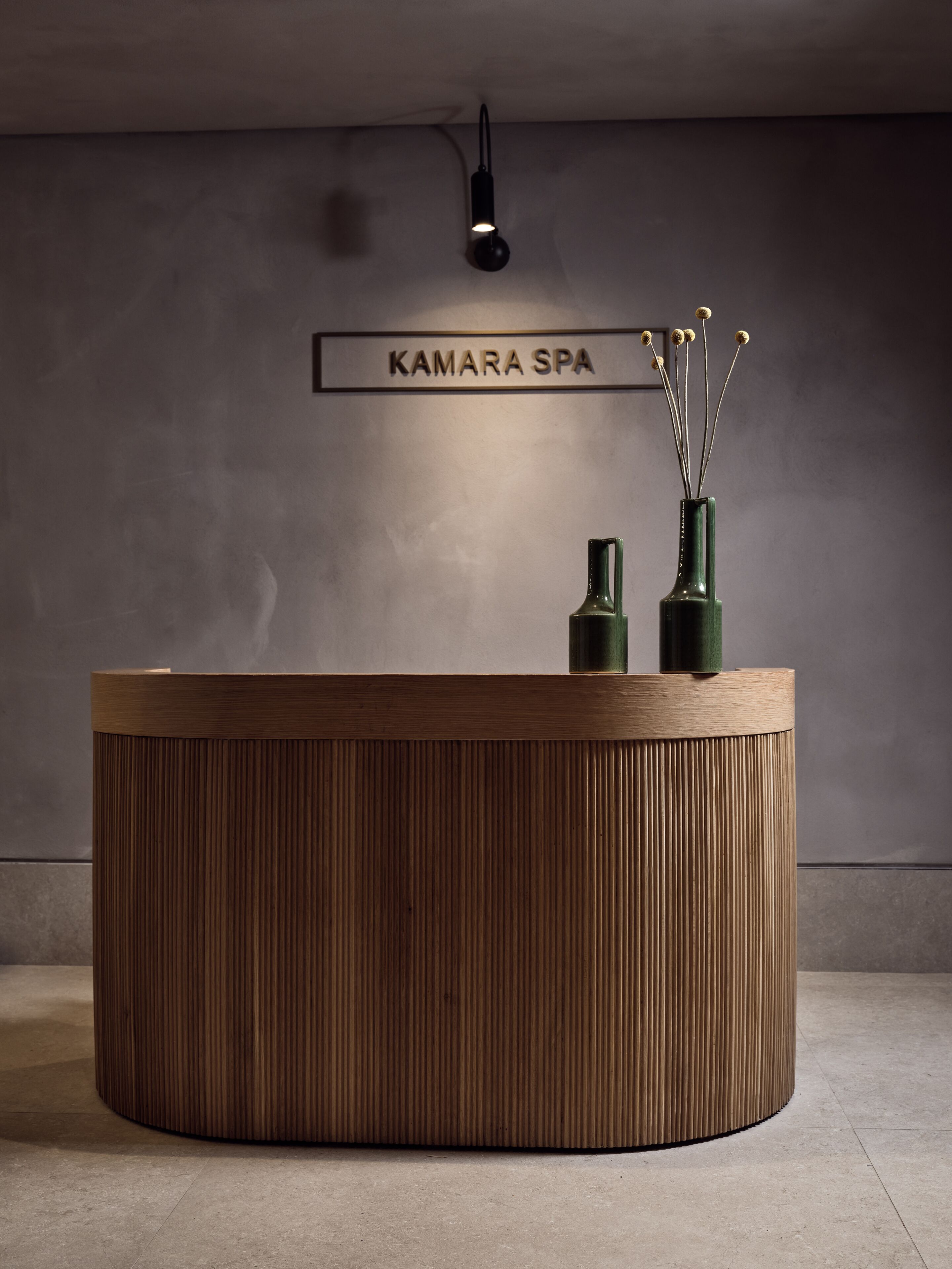 Ein Empfangsschalter mit Holzoptik, darüber ein Schild mit der Aufschrift "KAMARA SPA" und eine Vase mit getrockneten Blumen darauf.