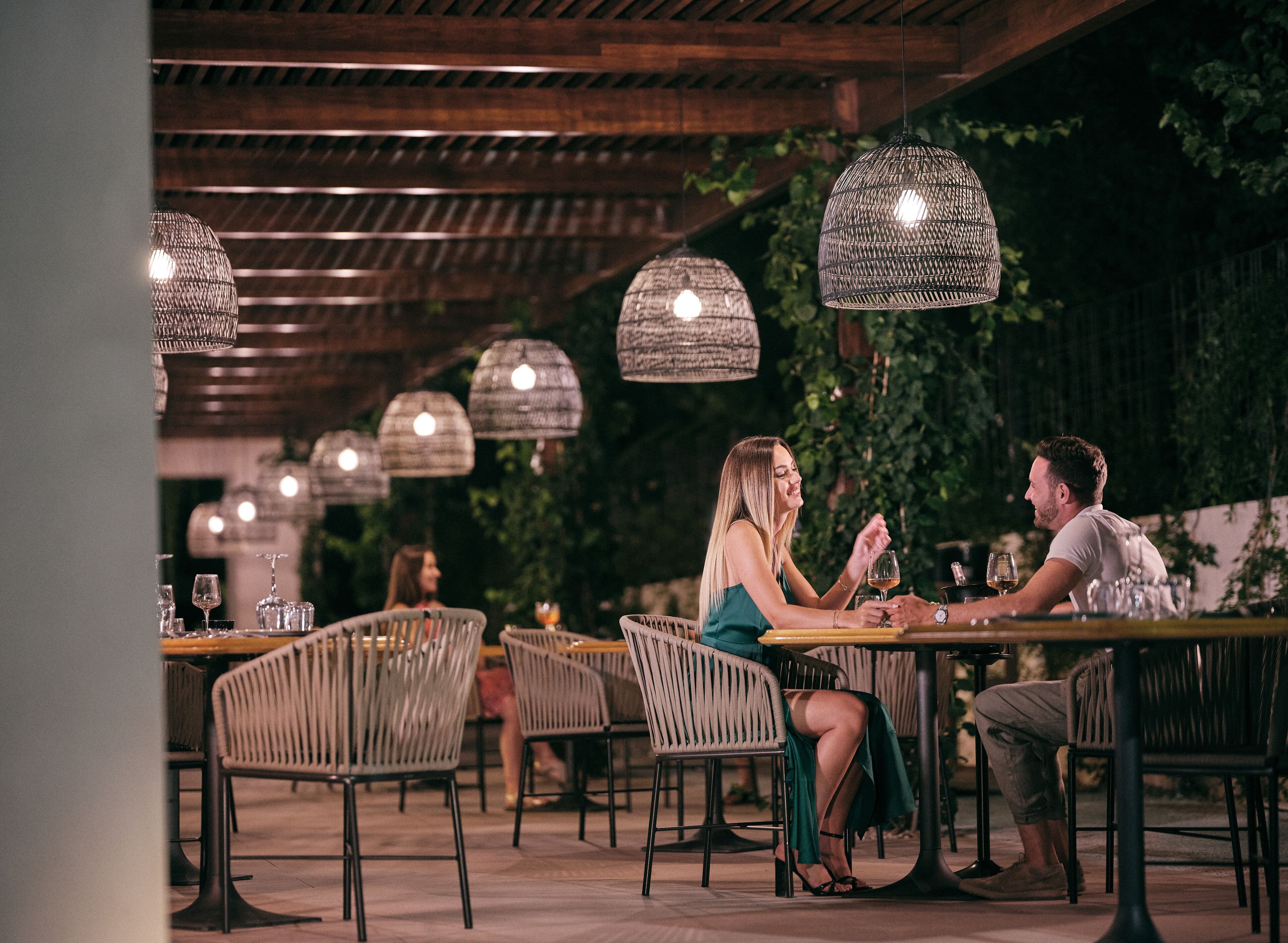 Un área de comedor al aire libre con sillas y mesas de mimbre, bajo un techo de madera con luces colgantes.