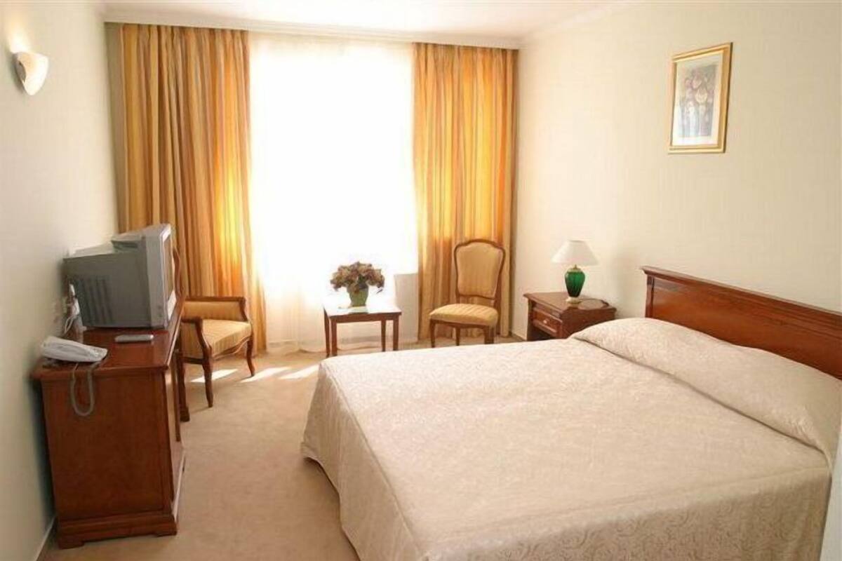 A hotel room with a bed, a desk with a lamp, a chair, a television, and a window with curtains.