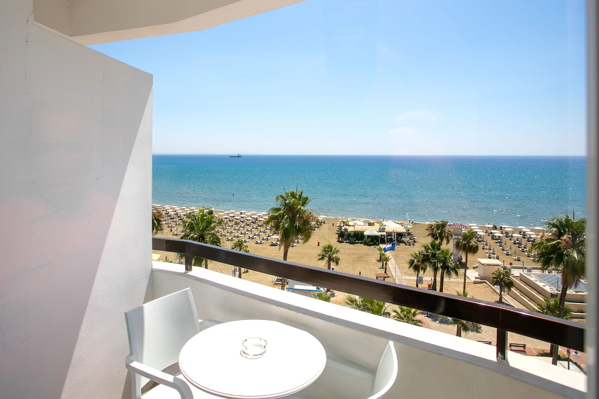 Un balcón con una mesa y sillas blancas, con vista a una playa con sombrillas y palmeras.