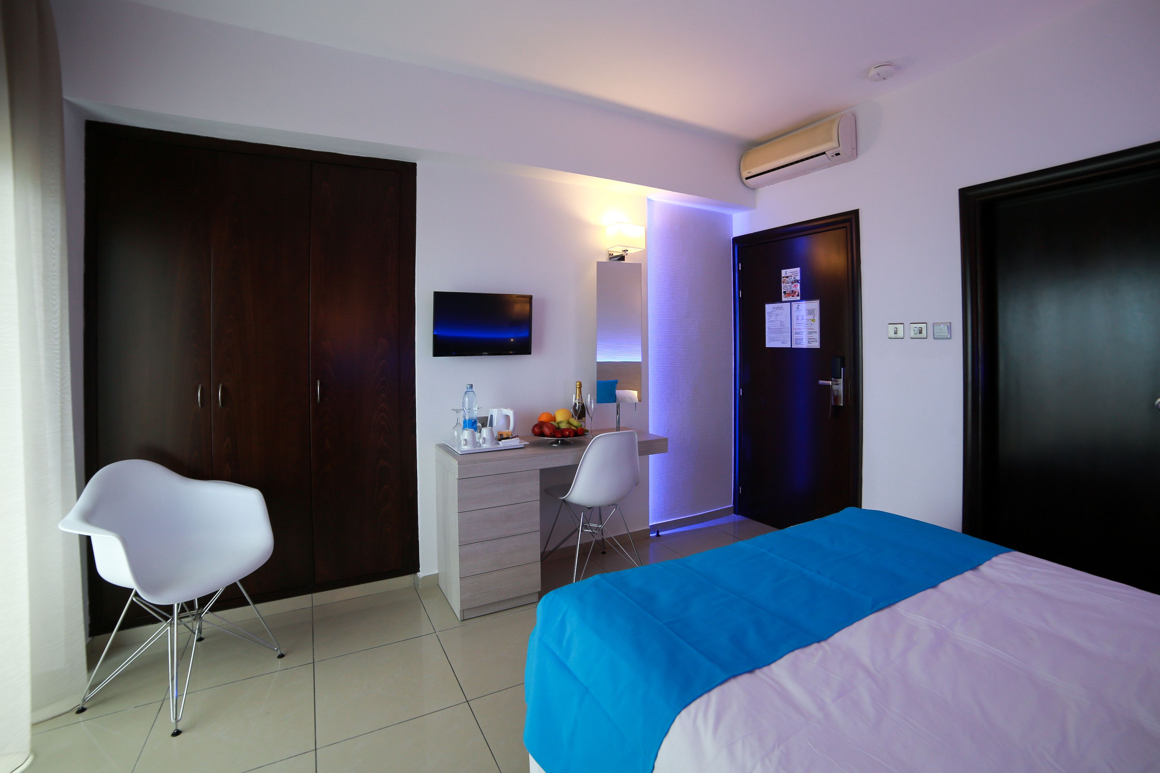 A hotel room with a bed, a desk, a chair, a wardrobe, a TV, and a door with a lock.