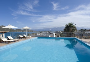 Zona de piscina con sillones de descanso y sombrillas, con vistas a un cuerpo de agua y montañas.