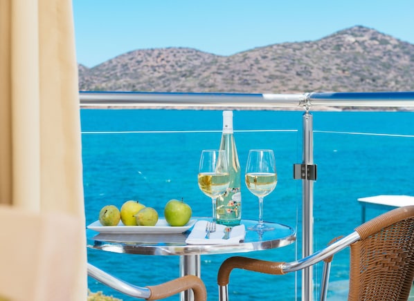 Una mesa con una botella de vino, dos copas y un plato de manzanas verdes en un balcón con vistas al mar.