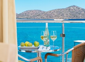 Una mesa con una botella de vino, dos copas y un plato de manzanas verdes en un balcón con vistas al mar.