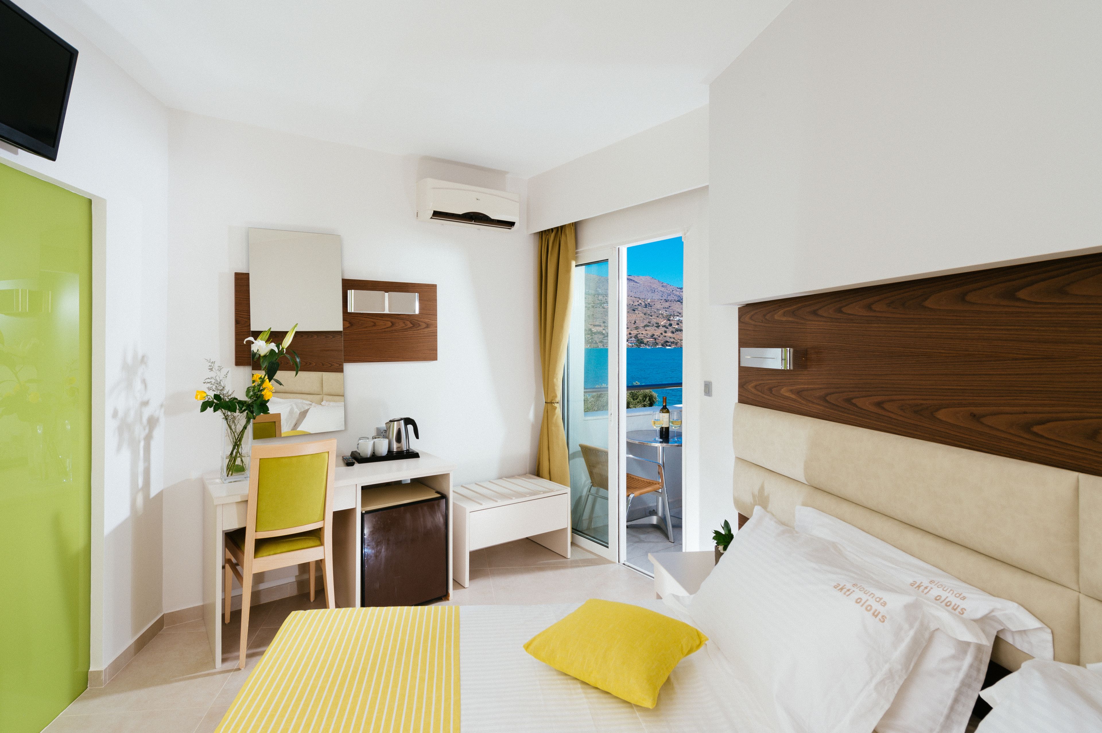 A modern hotel room with a bed, a desk, a chair, a small table, a mirror, and a view of the sea through a glass door.