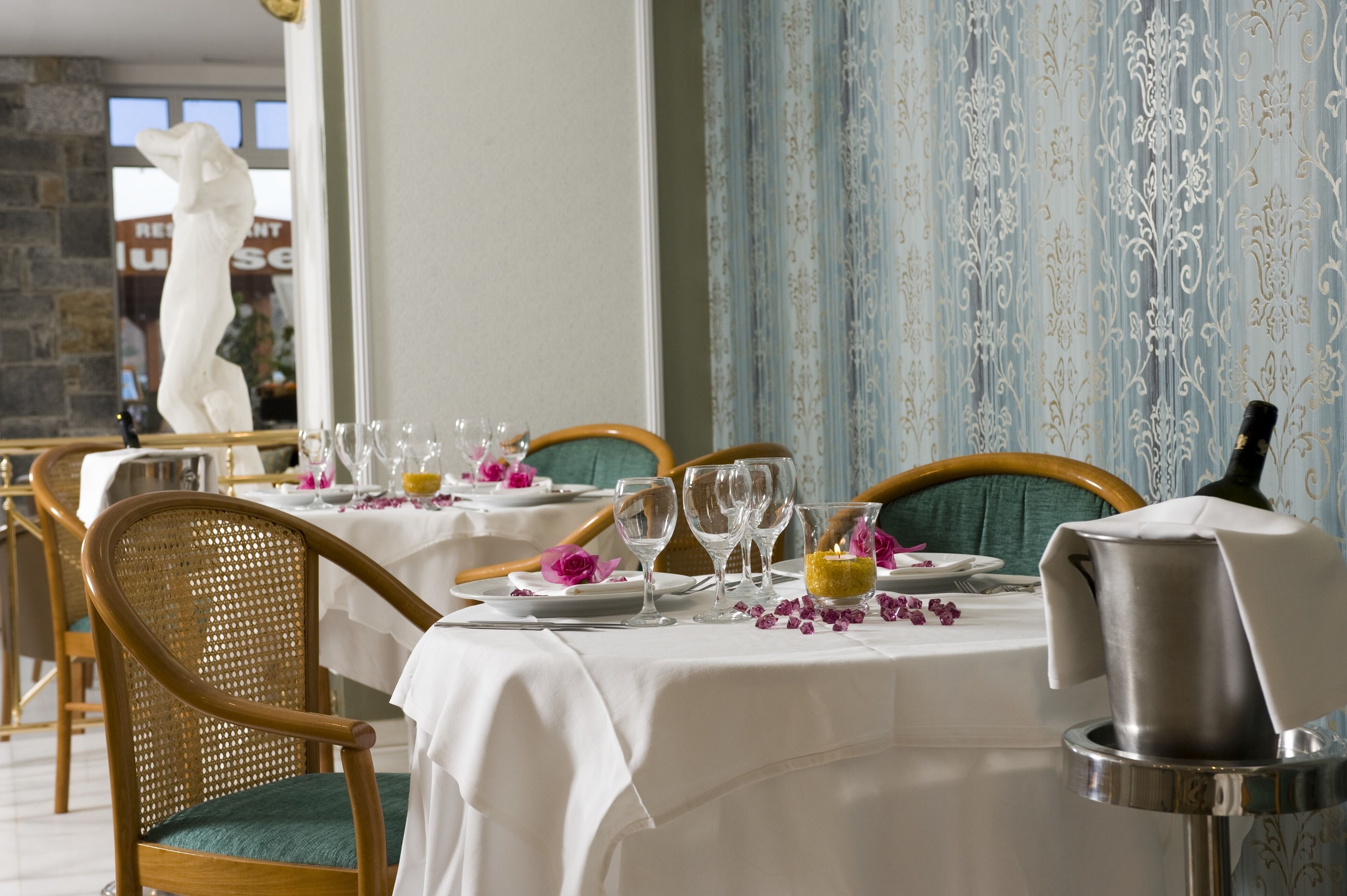 A dining area with tables set for a meal, featuring wicker chairs, white tablecloths, and a decorative centerpiece.