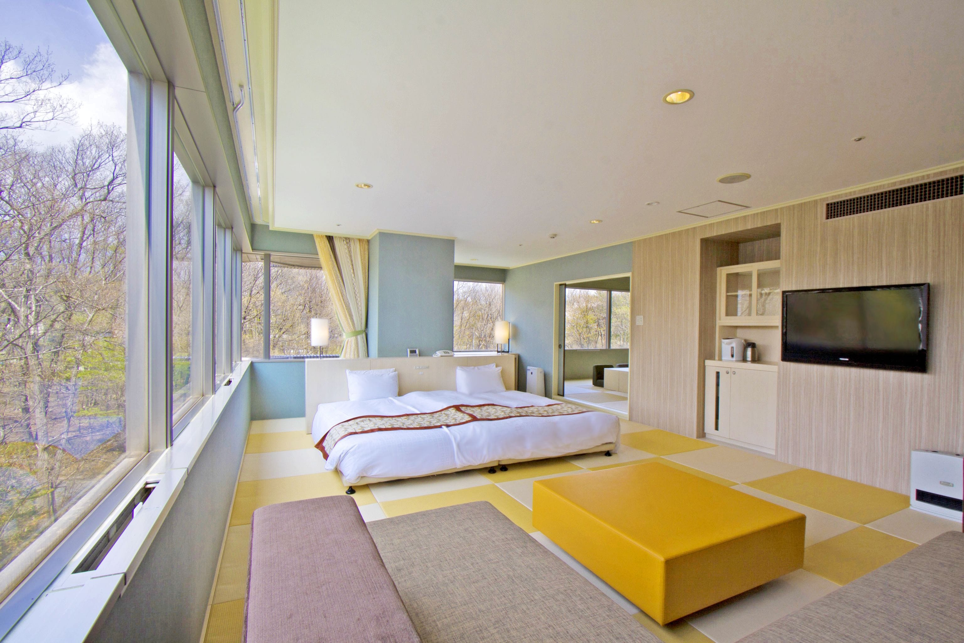 A modern bedroom with a large bed, a television, and a view of trees through large windows.