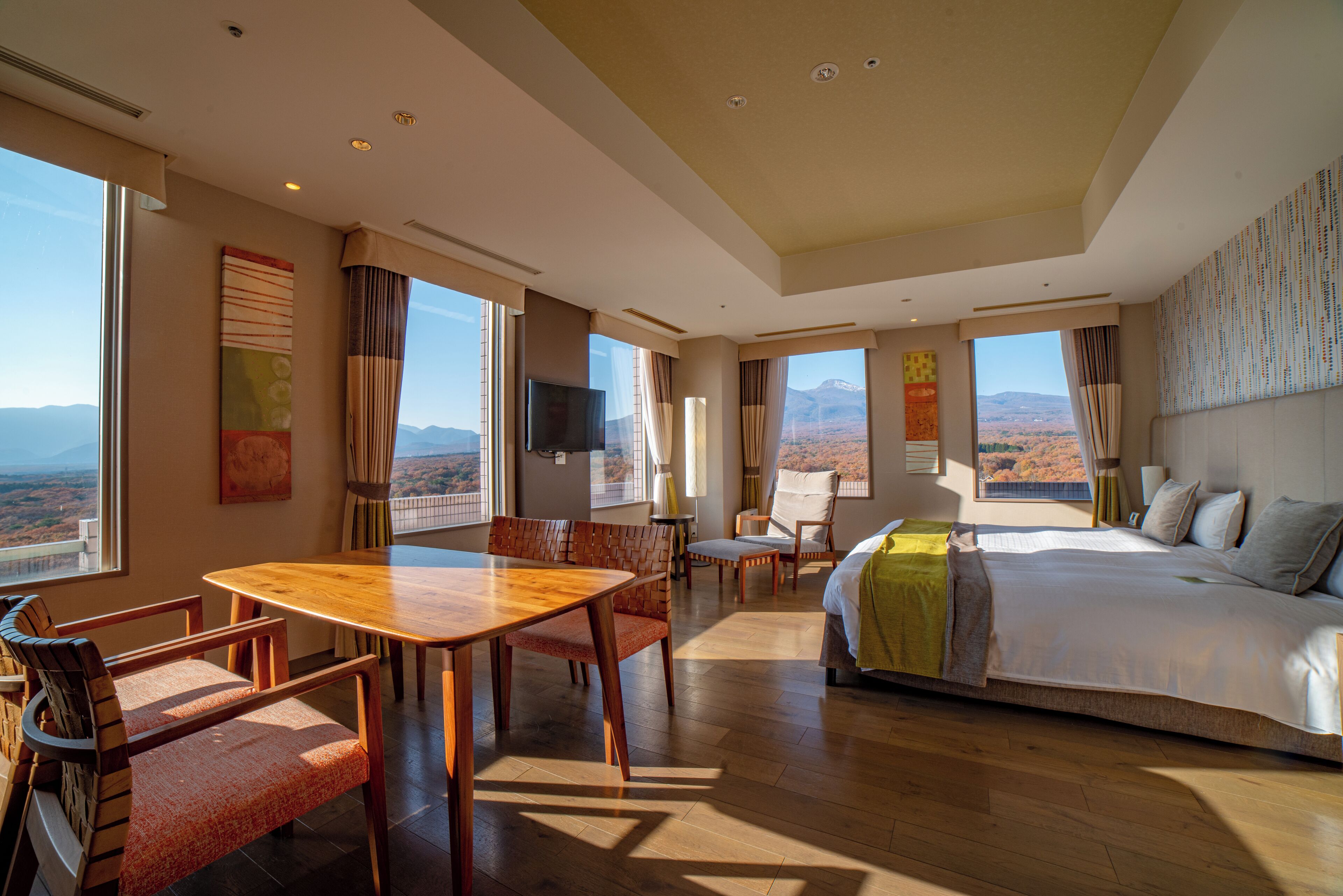 A hotel room with a large bed, a dining table, and a TV. There are multiple windows offering a view of the mountains.