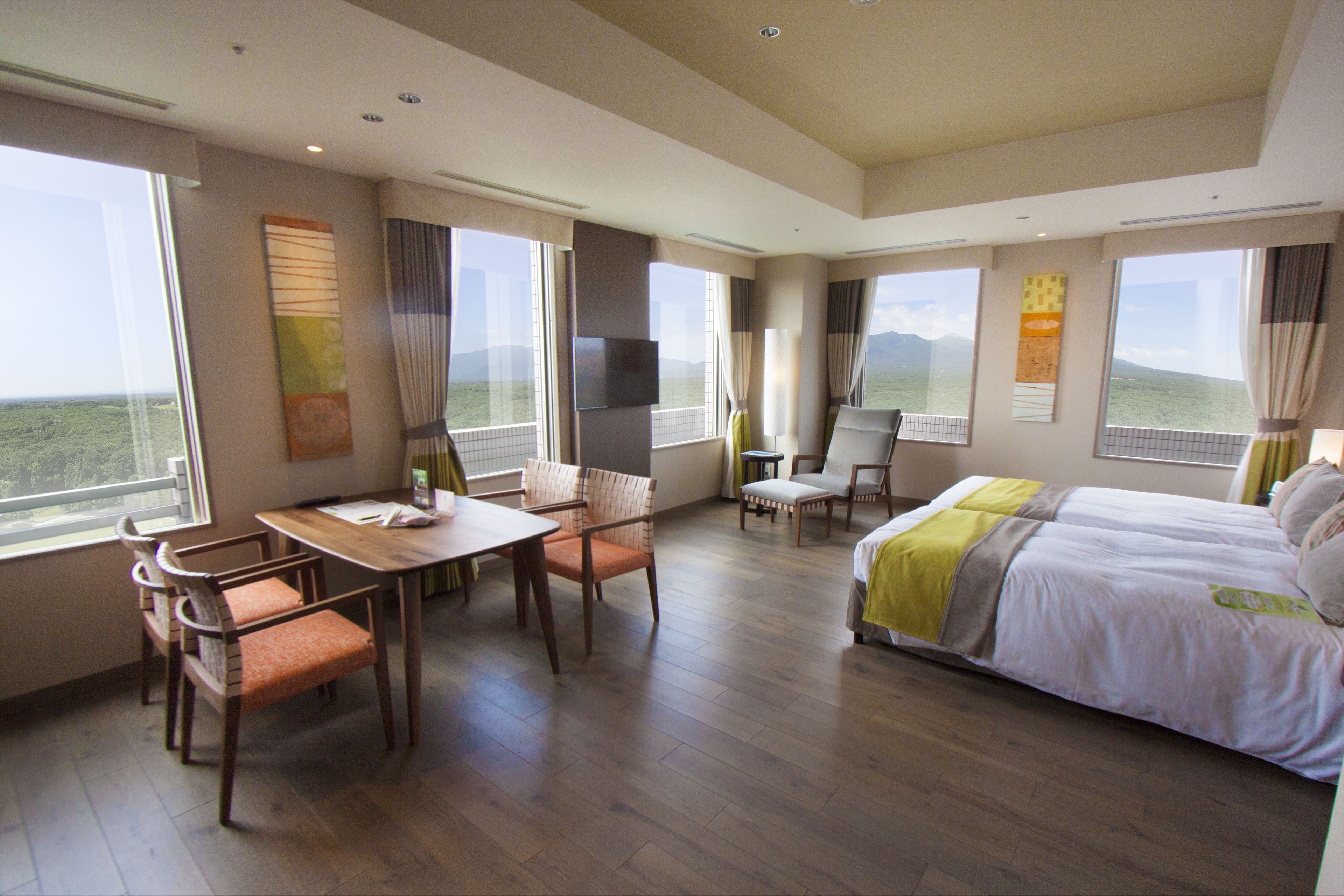 A hotel room with a bed, a dining table with chairs, a TV, and large windows offering a view of the landscape.