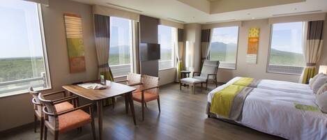 A hotel room with a bed, a dining table with chairs, a TV, and large windows offering a view of the landscape.