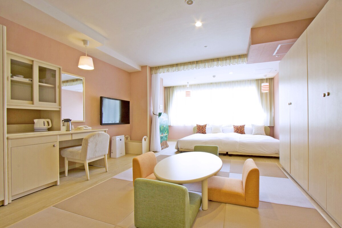 A hotel room with a bed, a desk with a chair, a television, and a window with curtains.
