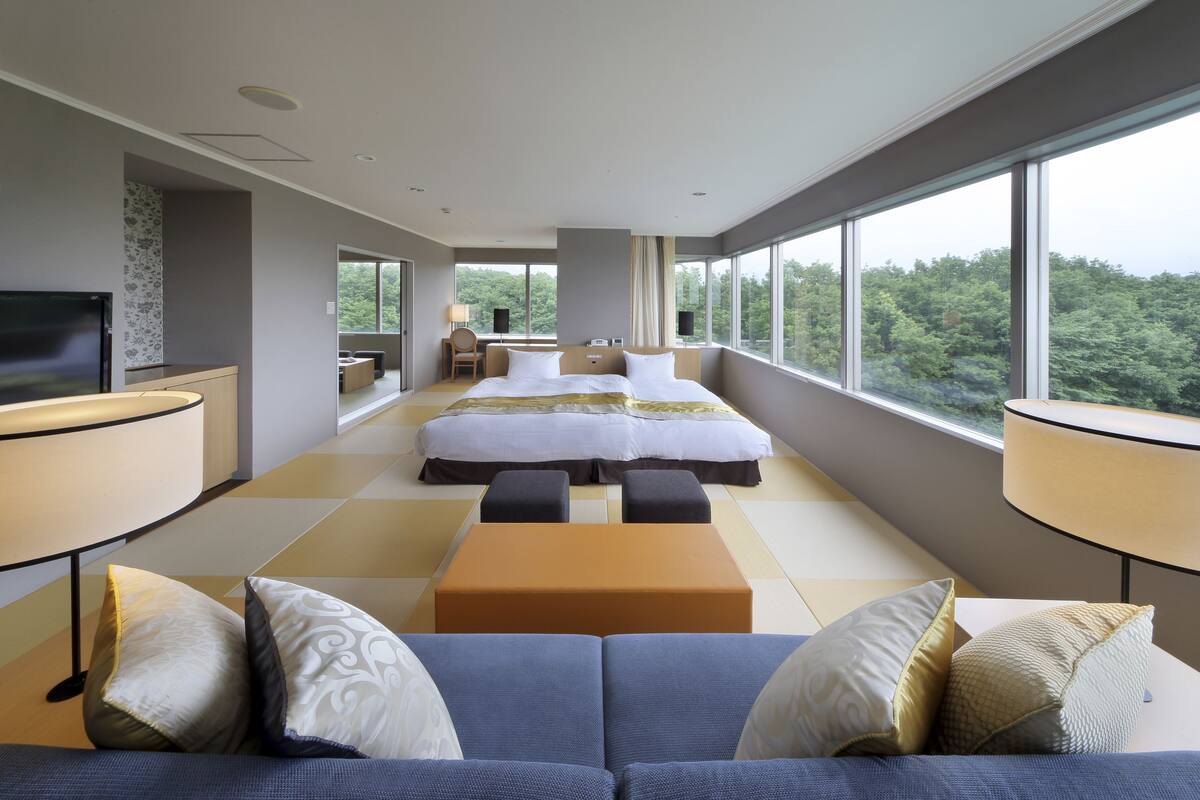 A modern hotel room with two beds, a sofa, a TV, and large windows offering a forest view.