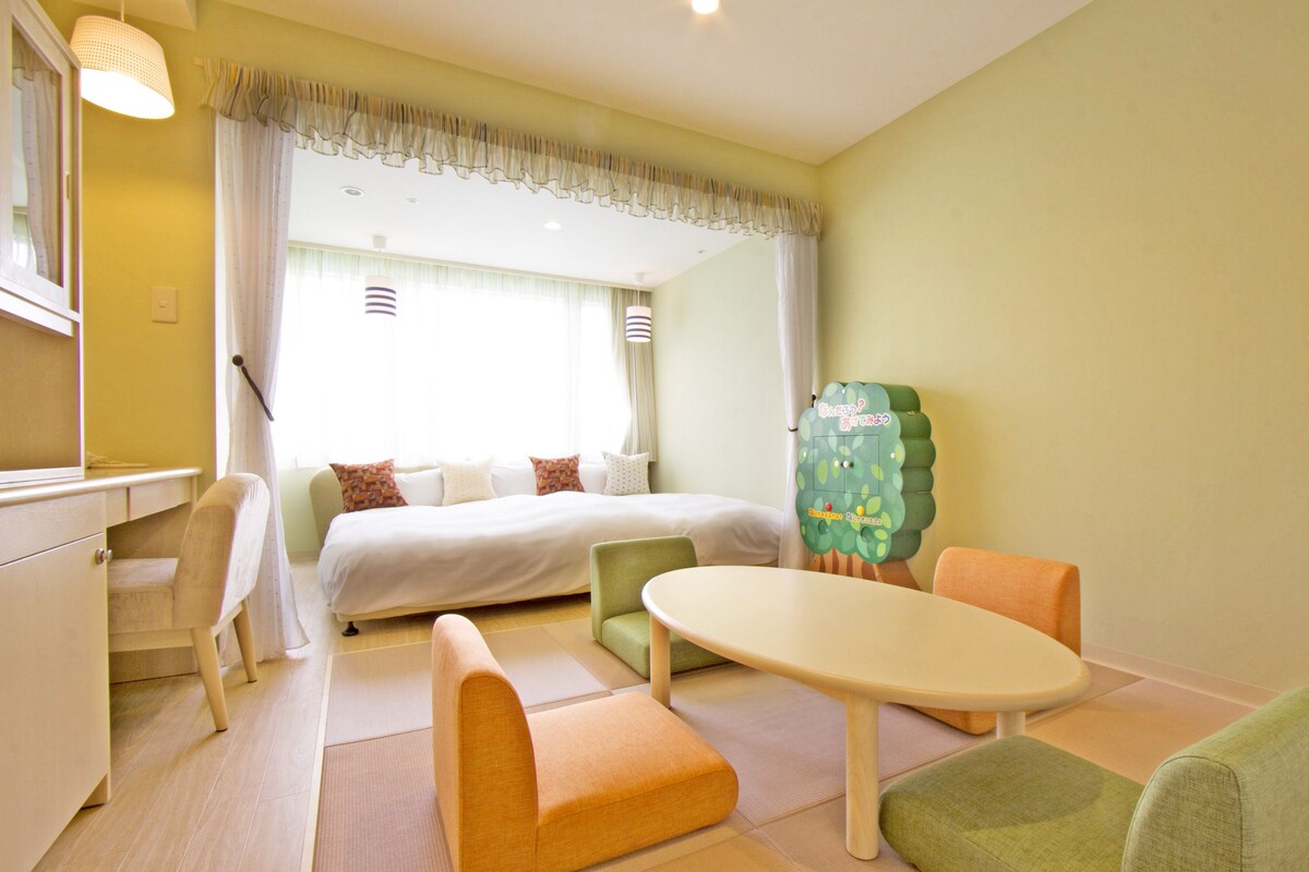 A hotel room with a bed, a desk, a chair, a table, and a green cabinet.