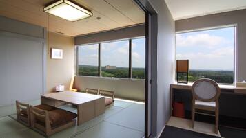A room with tatami mats, a low table, chairs, a bed, and a large window with a view of greenery.
