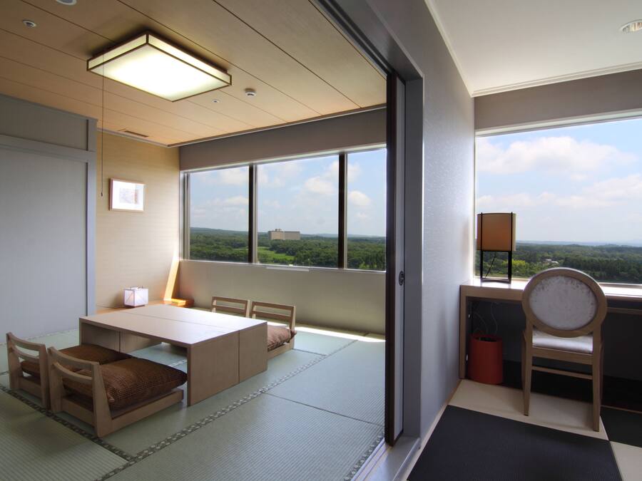A room with tatami mats, a low table, chairs, a bed, and a large window with a view of greenery.