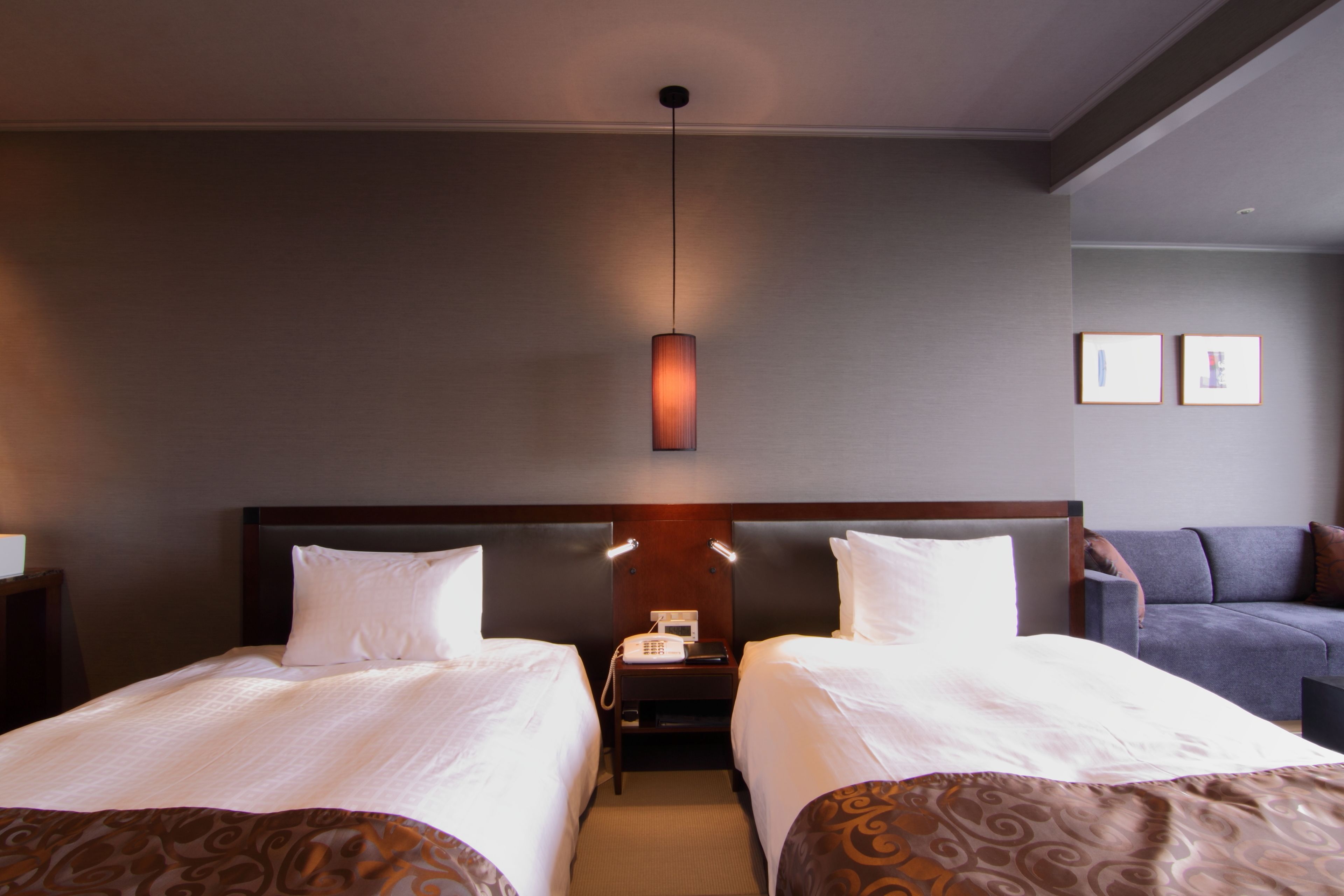 A hotel room with two beds, a bedside table with a phone, a wall-mounted lamp, and a sofa.