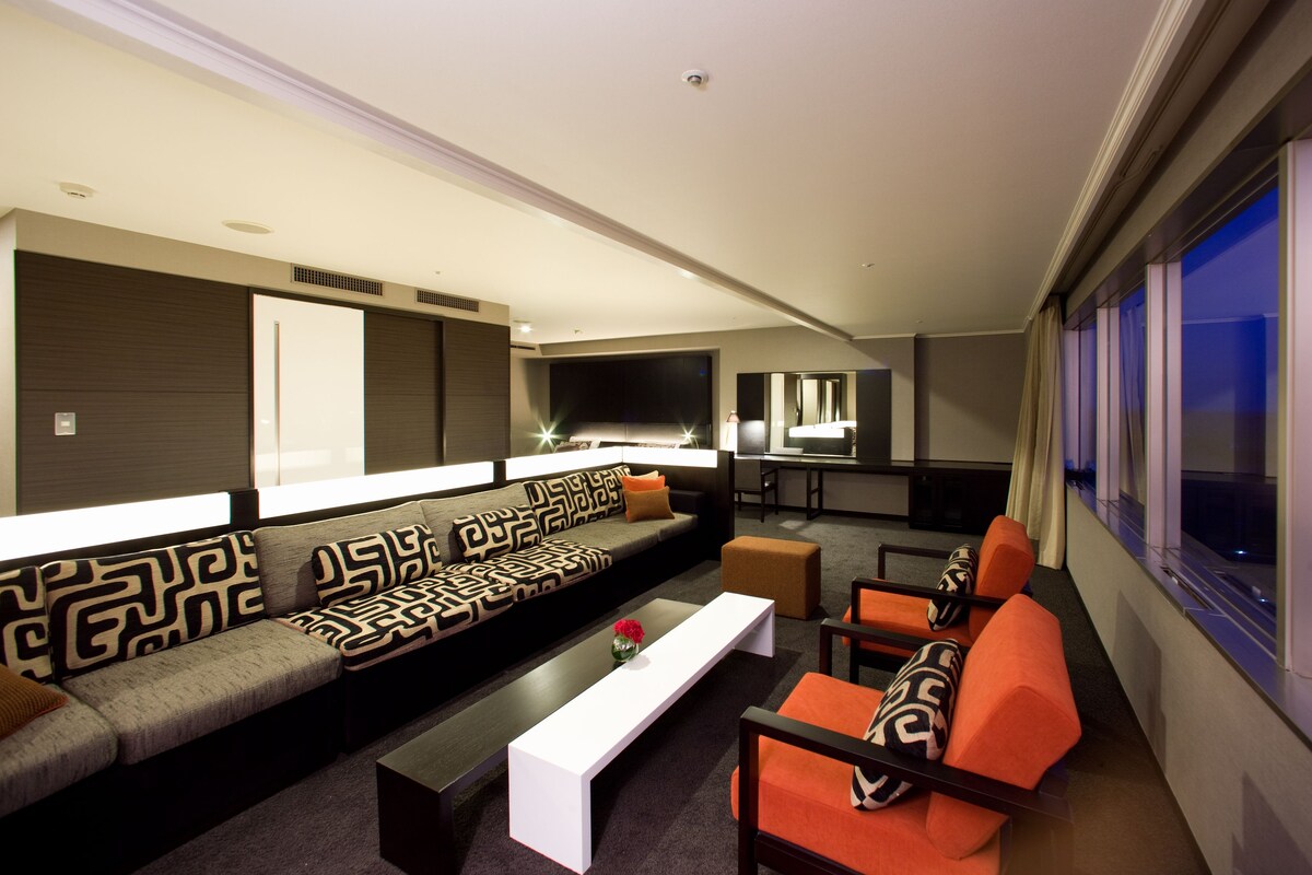 A modern living room with a large sectional sofa, a flat-screen TV, a coffee table, and a seating area with orange chairs.