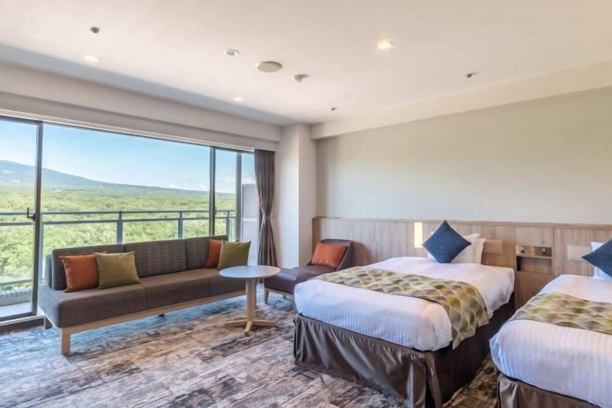 A hotel room with a large window, a sofa, two beds, and a small table.