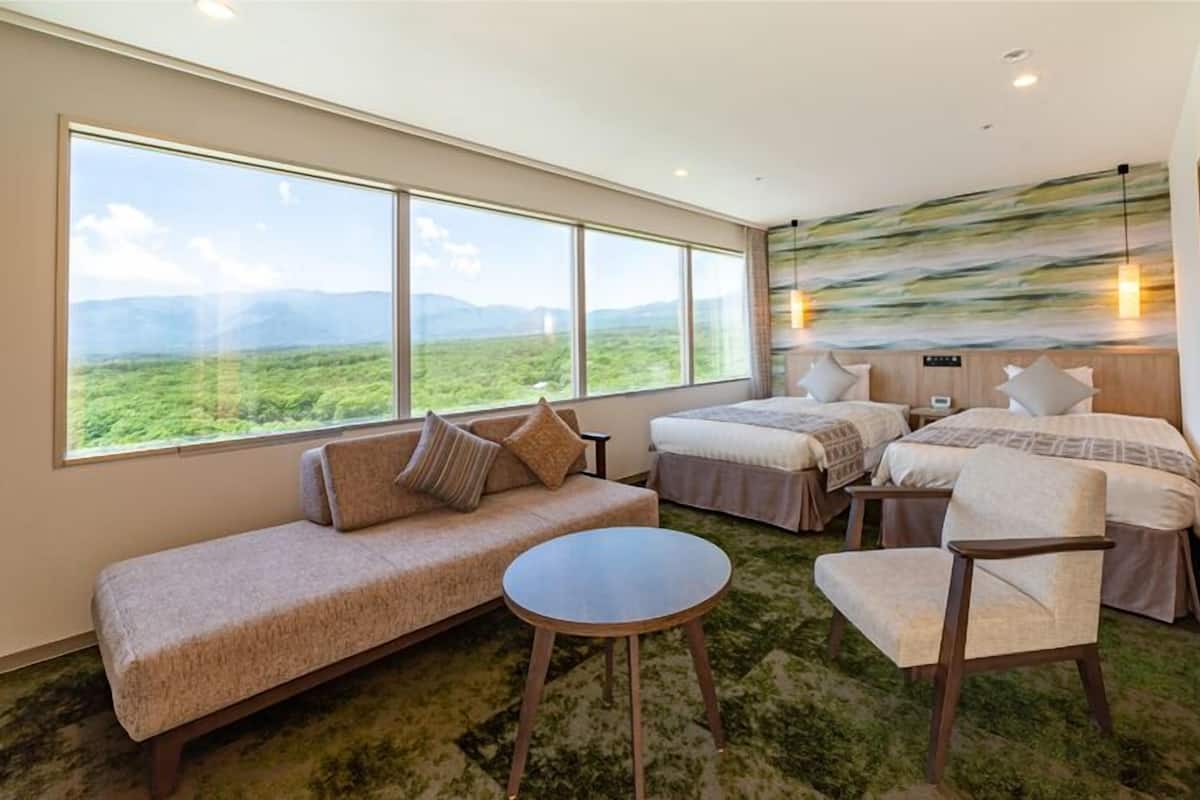 A hotel room with a large window, a sofa, two single beds, a round table, and a mural on the wall.