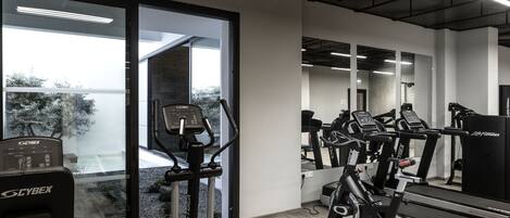 Une salle de sport équipée de machines Cybex, dotée de grandes fenêtres et offrant une vue sur les arbres.