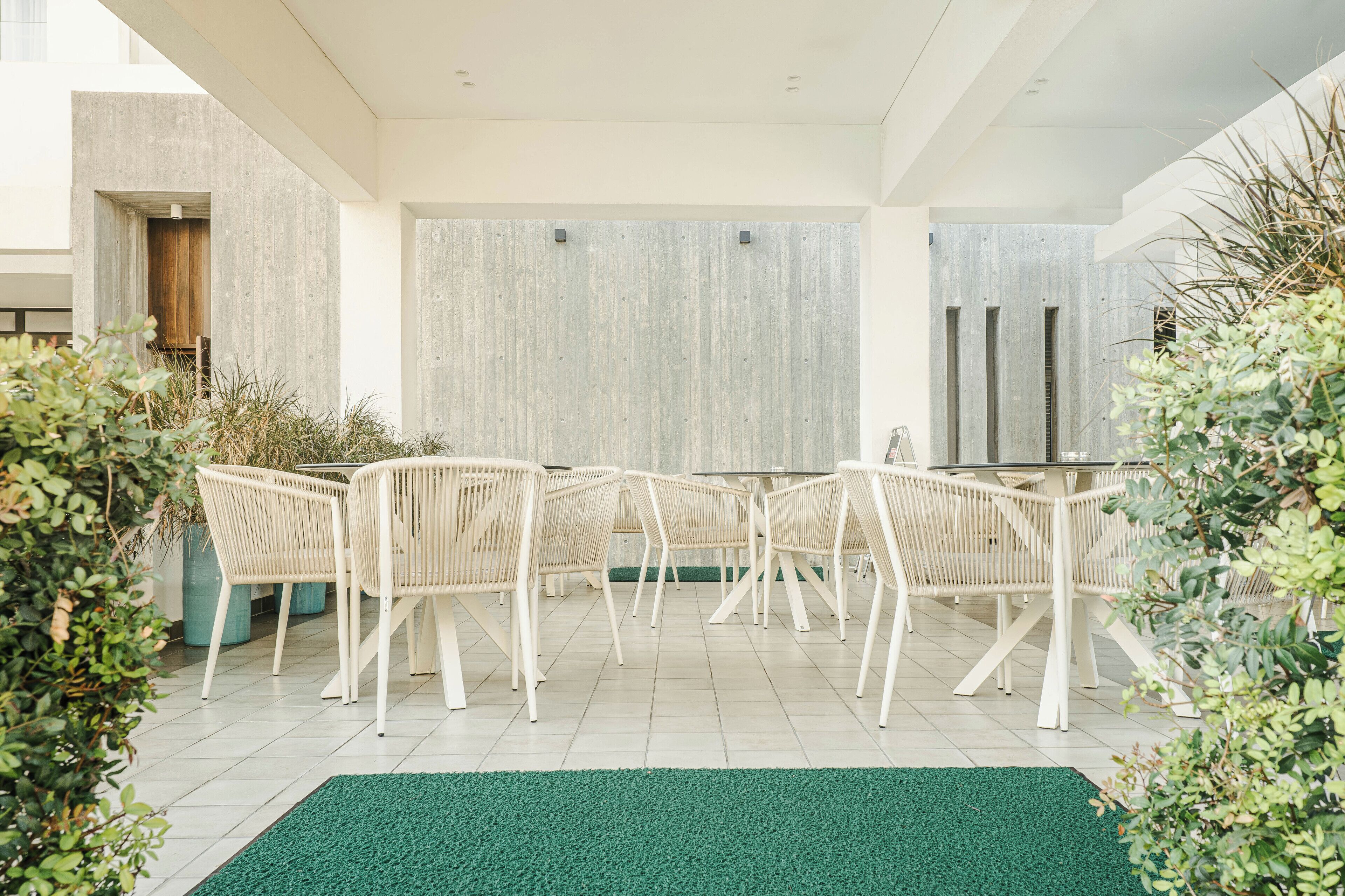 A modern outdoor dining area with white wicker chairs and a glass table, surrounded by greenery and a green artificial turf floor.