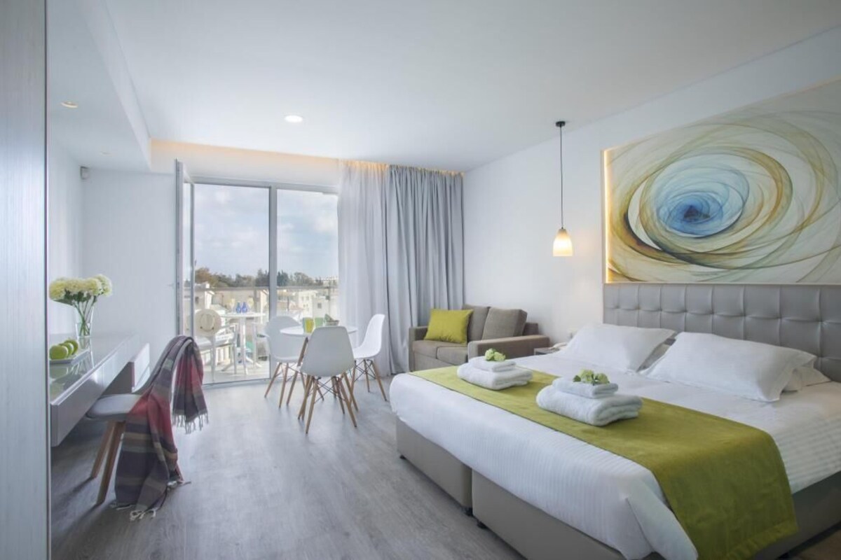 A modern hotel room with a large bed, a desk with a chair, a balcony view, and a large abstract painting on the wall.
