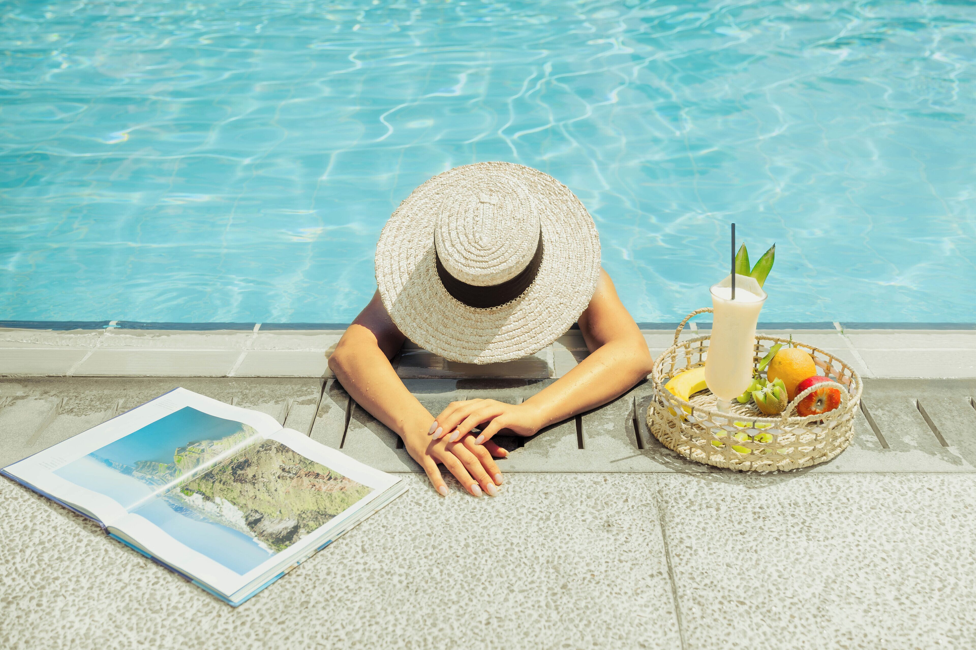 Une personne assise au bord d’une piscine, avec une boisson servie dans un verre muni d’une paille, un panier de fruits et un magazine.