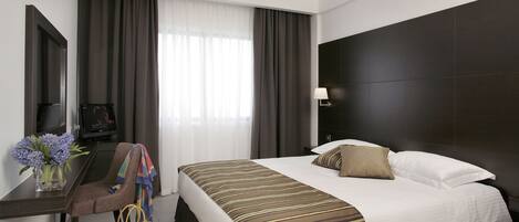 A hotel room with a bed, a desk with a chair, a TV, and a lamp.