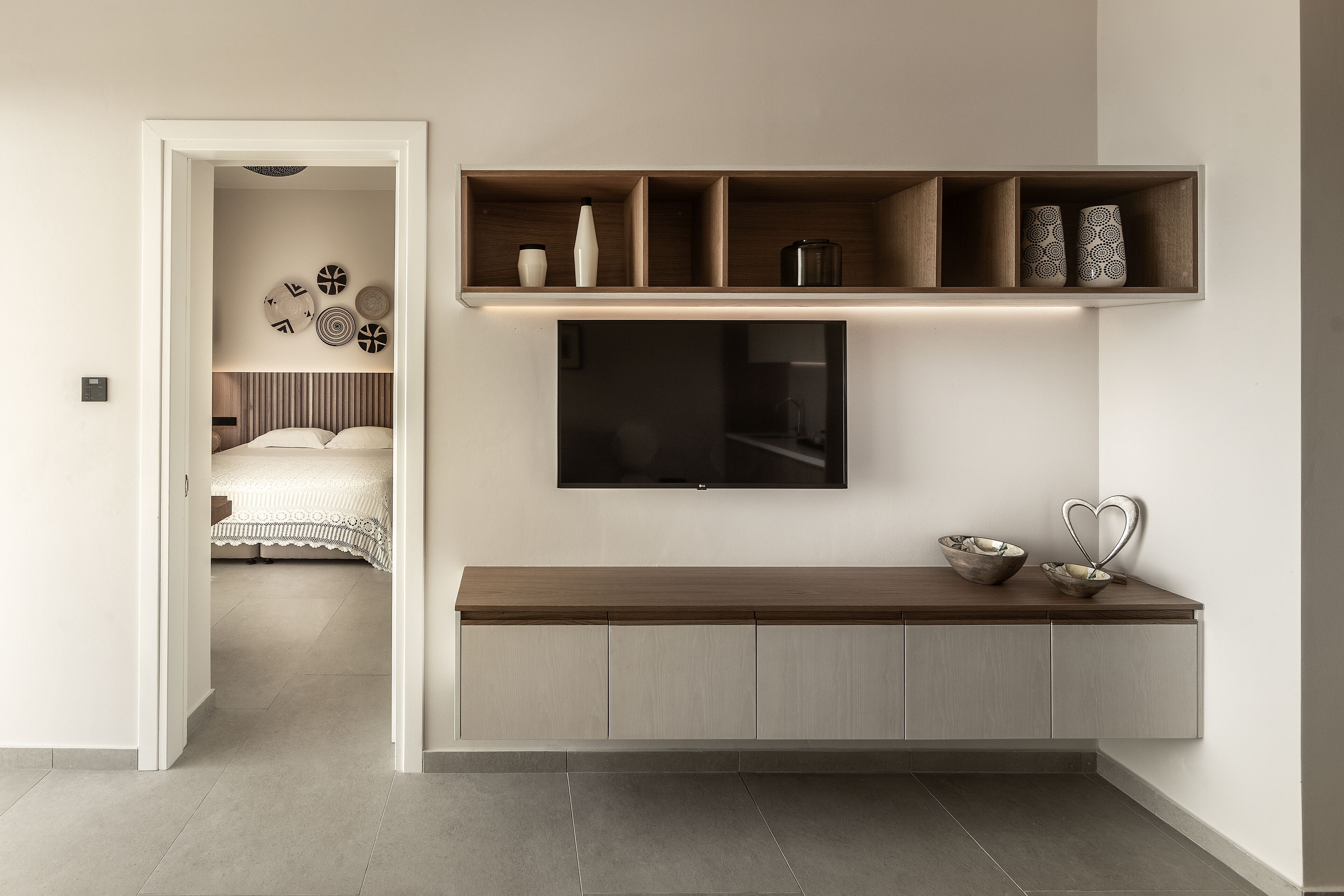 Quarto moderno com cama, televisão fixada na parede e mesa consola com objetos decorativos.