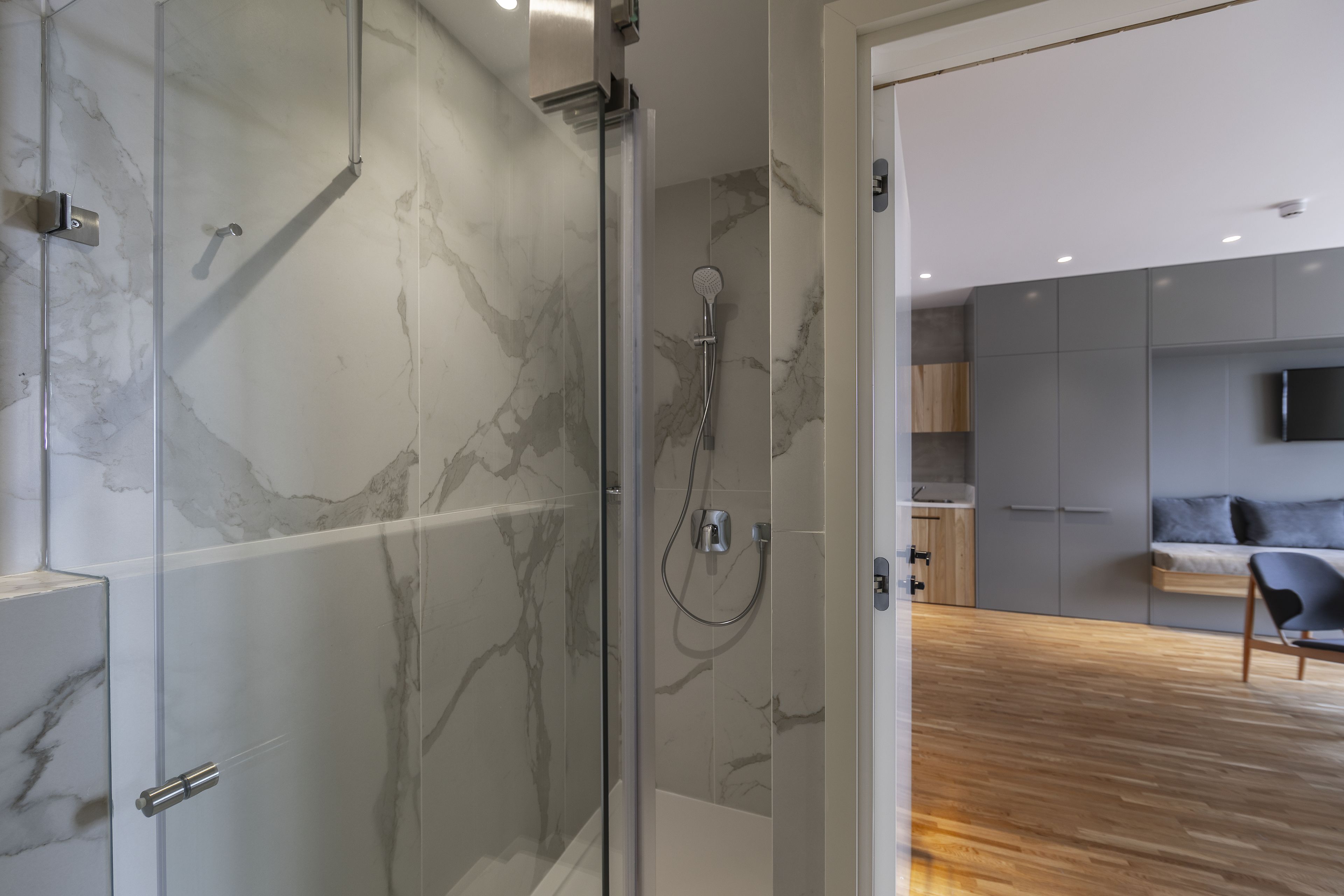 A modern bathroom with a glass-enclosed shower, marble walls, and a wooden floor.