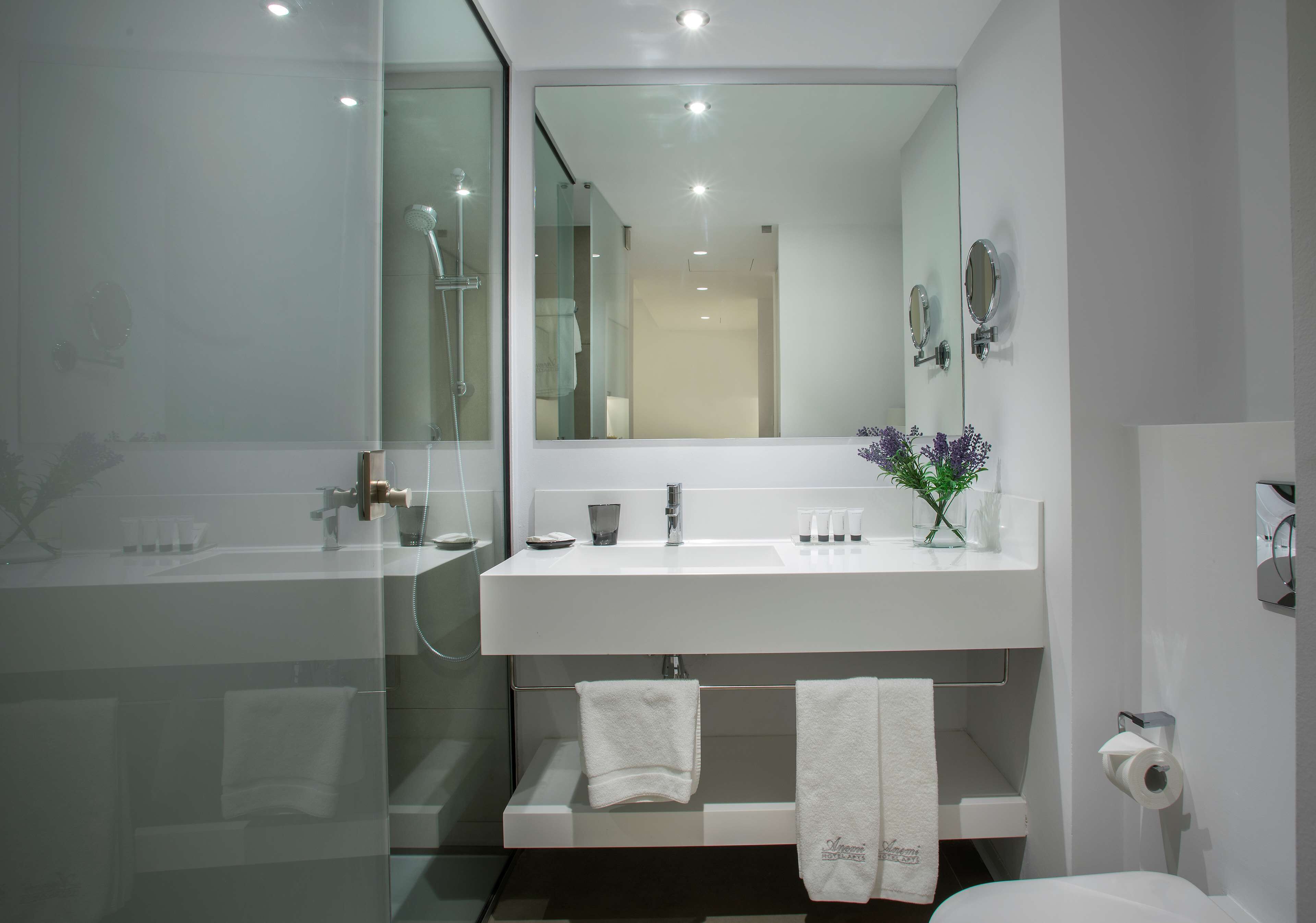 A modern bathroom with a glass-enclosed shower, a white sink with a mirror above it, and a toilet with a roll of toilet paper on the wall.