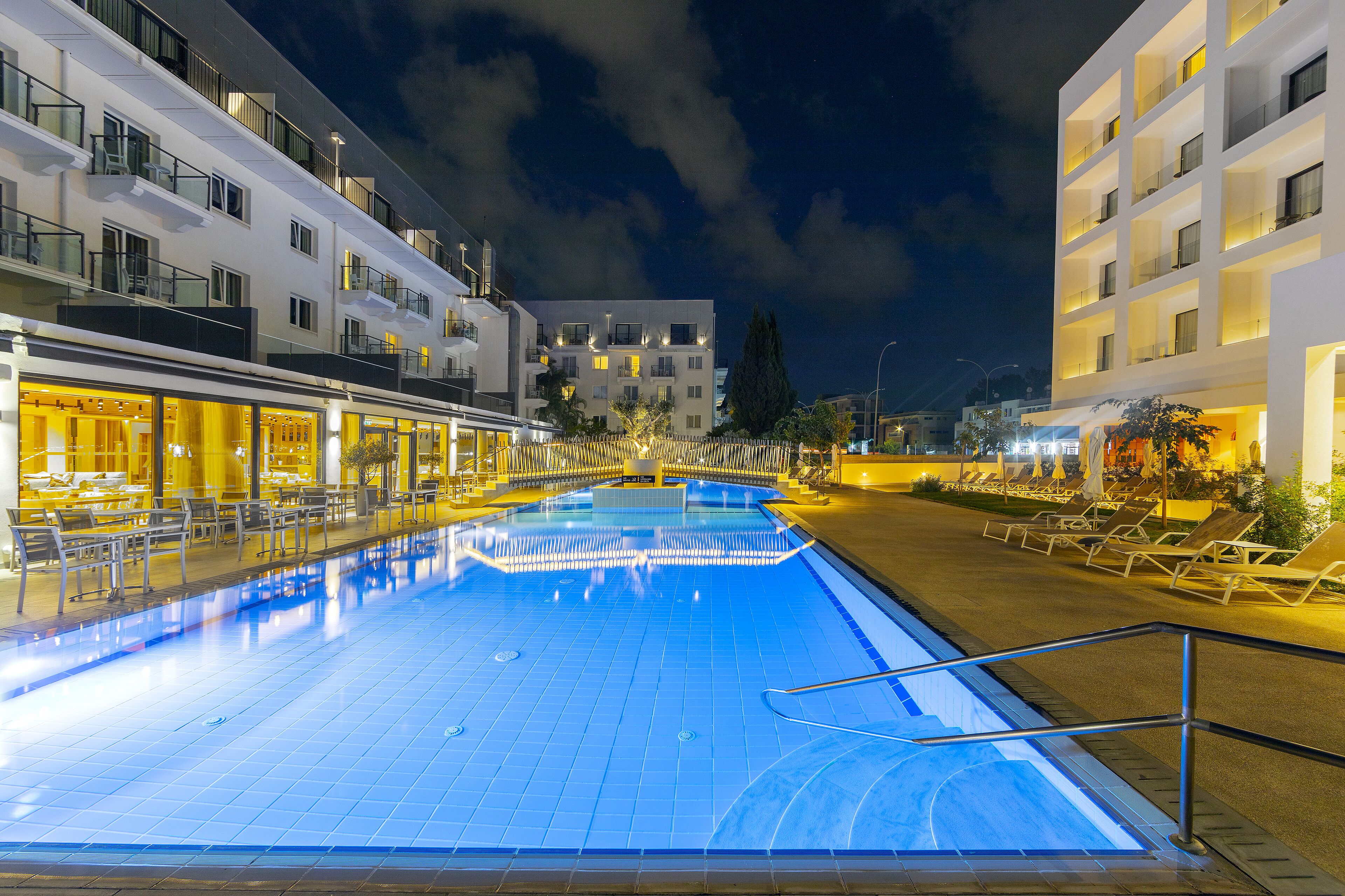 A nighttime swimming pool with adjacent lounge areas and a building with multiple balconies.