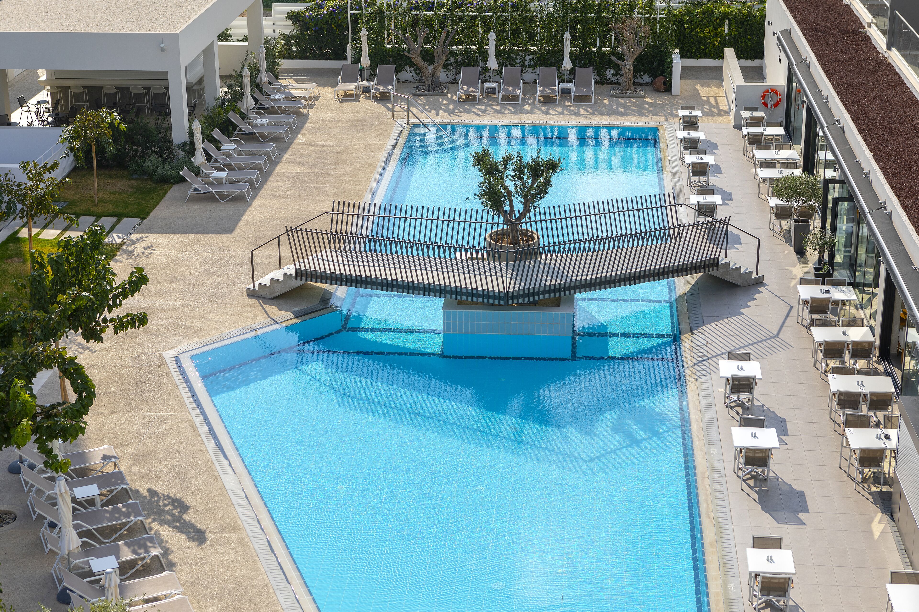 A large rectangular swimming pool with a central island, surrounded by lounge chairs and tables, and a covered area with umbrellas.