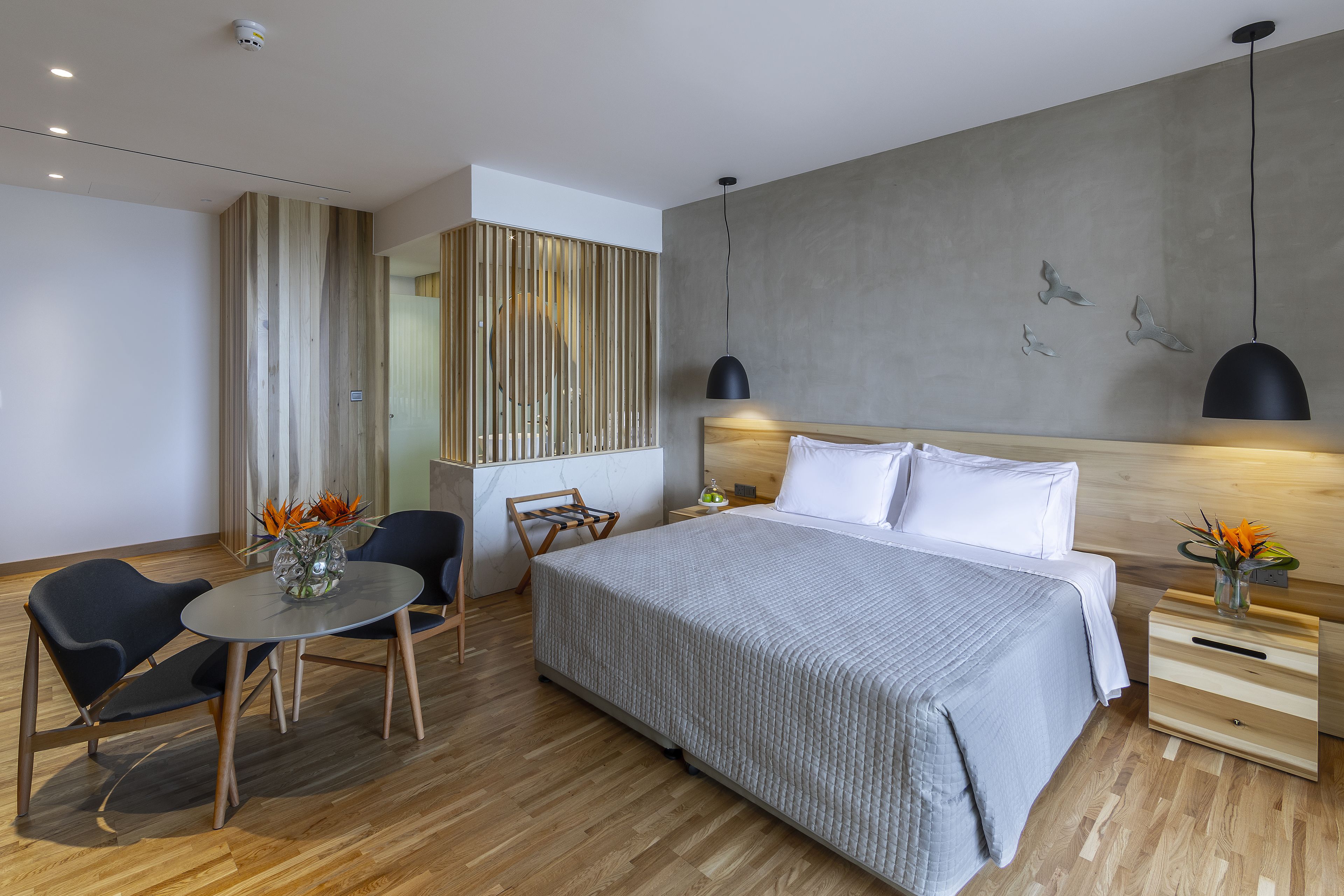 A modern hotel room with a large bed, a wooden floor, a dining table with chairs, and a small kitchen area.