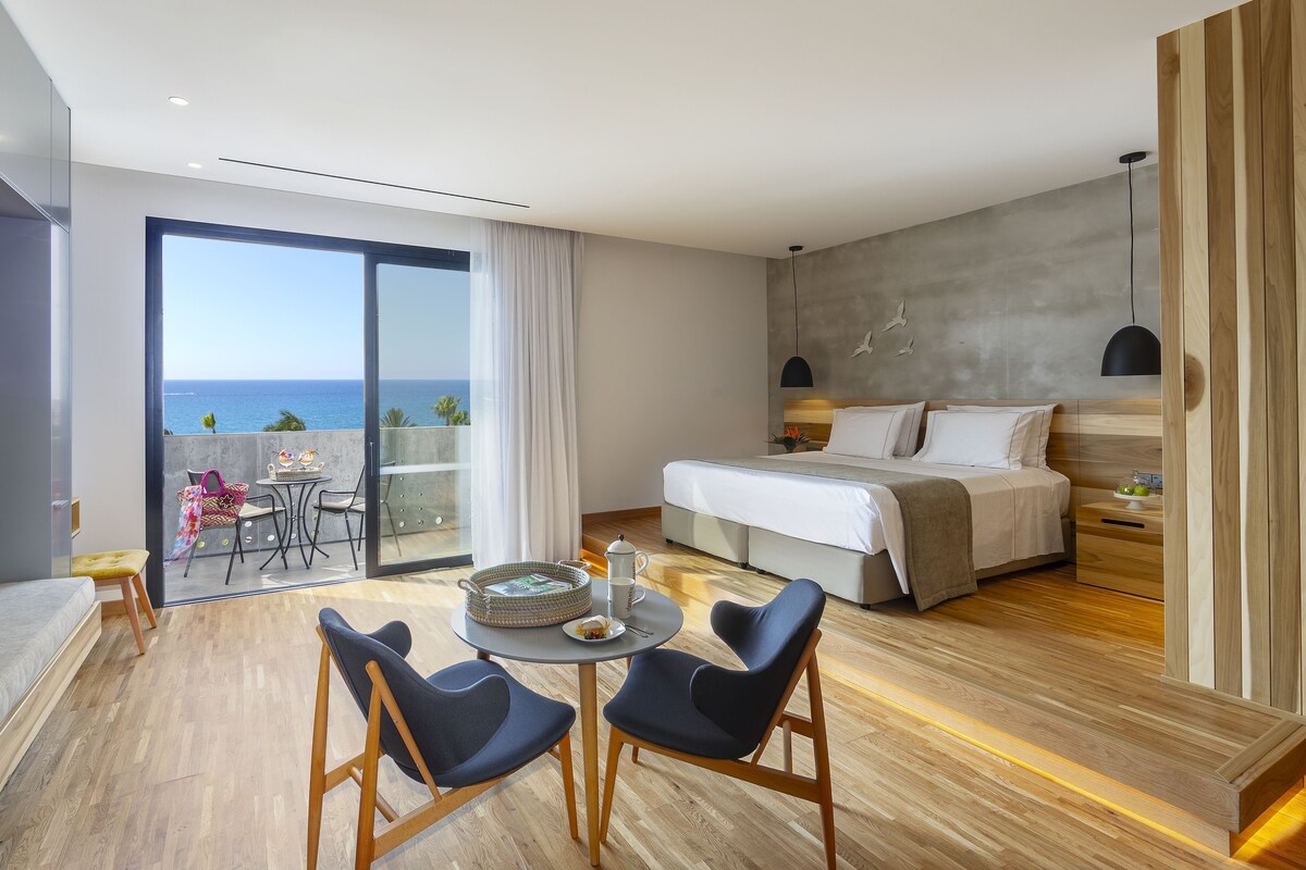 A modern hotel room with a balcony, a bed, a dining area, and a view of the sea.